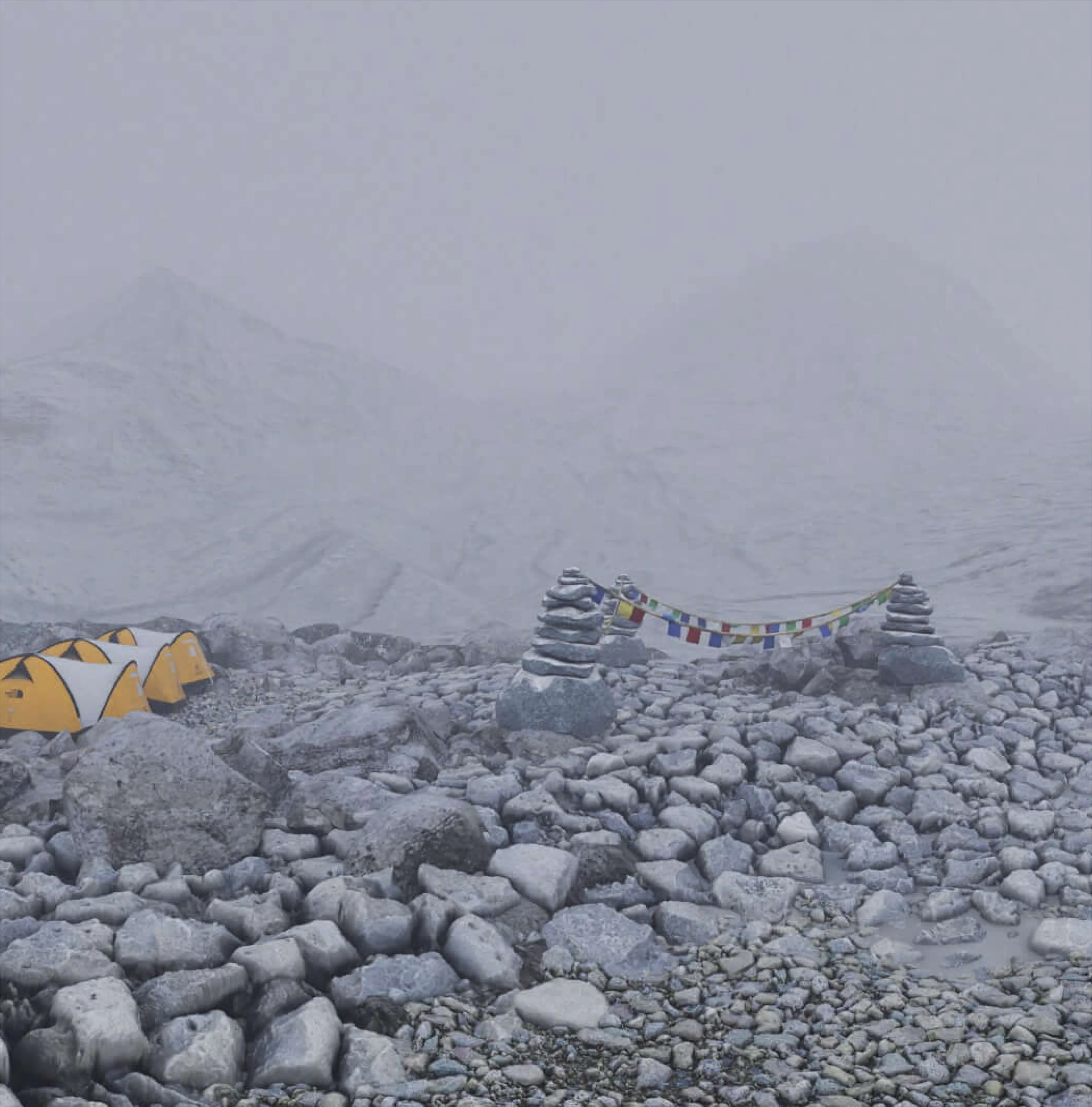 Base camp shrouded in fog: prayer flags, stone cairns, and a cluster of orange tents on the boulder field.