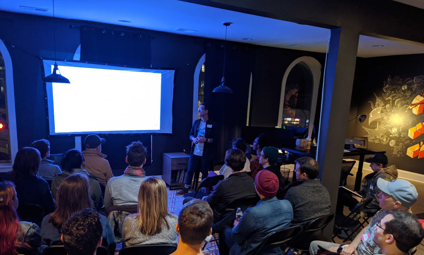A speaker presenting to a seated audience in a dimly lit room at a Rough Draft session, a lit projector screen behind him