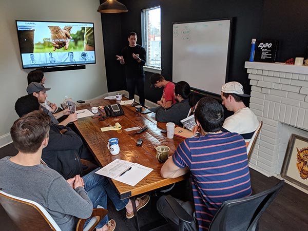 A small group gathered around a wooden table watching a presenter at a screen in the Triptych office